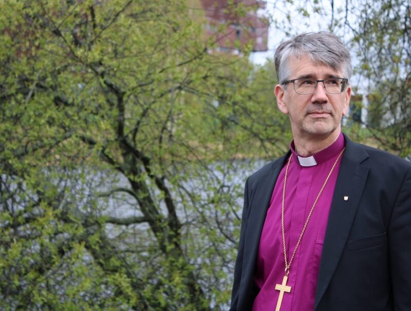 Biskopen är klädd i lila biskopsskjorta och svart kavaj, han har grått hår och glasögon och står utomhus framför ljusgrönt grönskande träd.