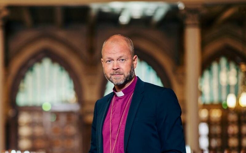 Biskopen är klädd i lila biskopsskjorta och mörkblå kavaj, han har kortklippt hår och skägg och står inne i en kyrka med gotiska fönster.