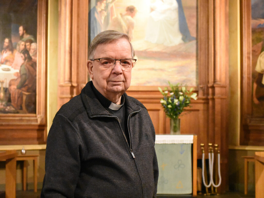 En äldre man i prästskjorta, grå tröja och glasögon står inne i en kyrka med många målningar.