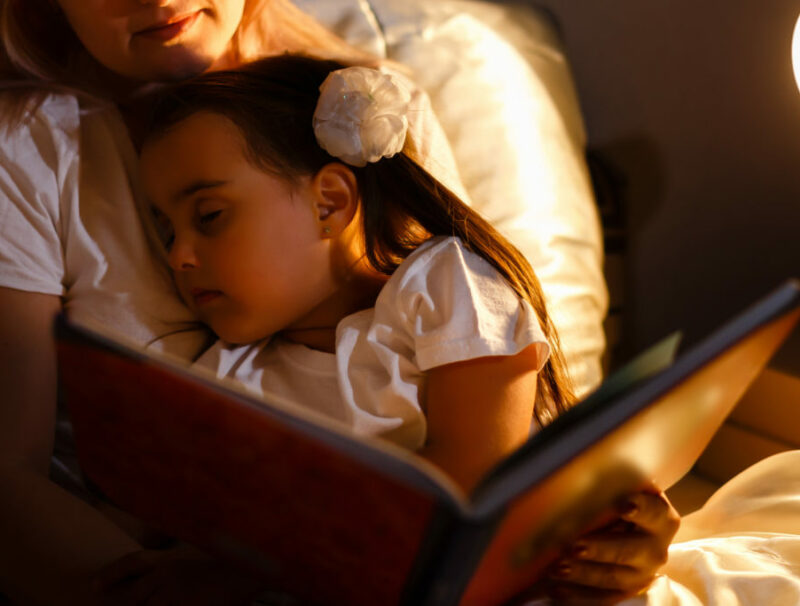 En liten flicka sover i famnen på en vuxen som läser en bok för henne.