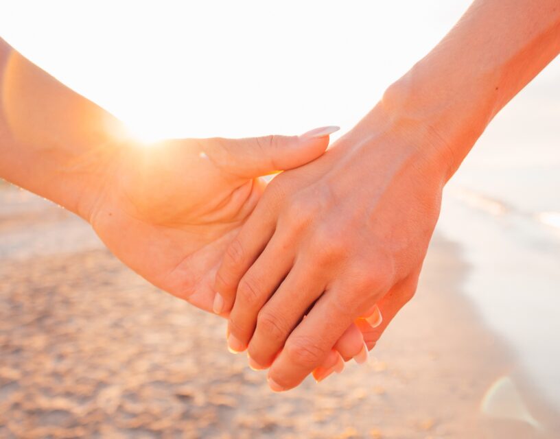 Pari pitää toisiaan kädestä. Taustalla hiekkaranta ja auringonvaloa.