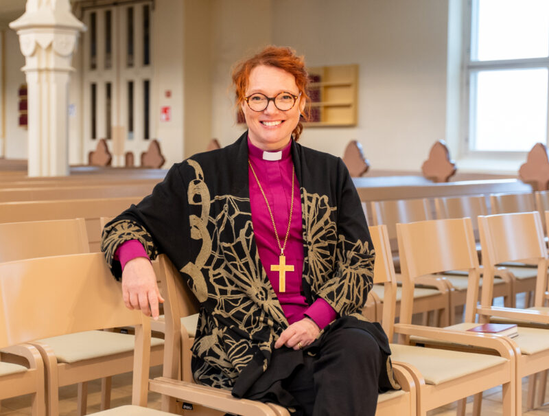 Biskopen är klädd i lila biskopsskjorta och en svart och guldfärgad kofta, hon har rött, lockigt hår och glasögon och sitter i en stolsrad i en kyrksal.