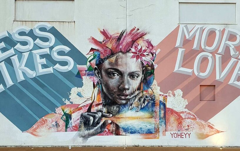 En väggmural föreställande en ung kvinna omgiven av färger och blommor och texten "Less Likes – More Love".