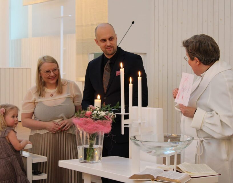 Frida och Samuel samt en liten flicka står vid dopfunten med en präst.