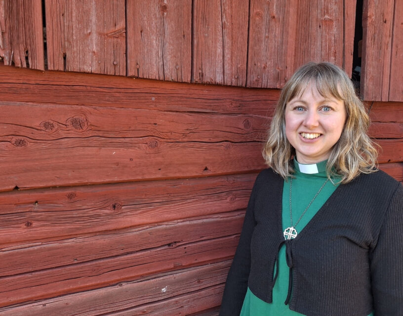 Jessica Emaus i grön diakonskjorta framför en rödmålad trävägg.